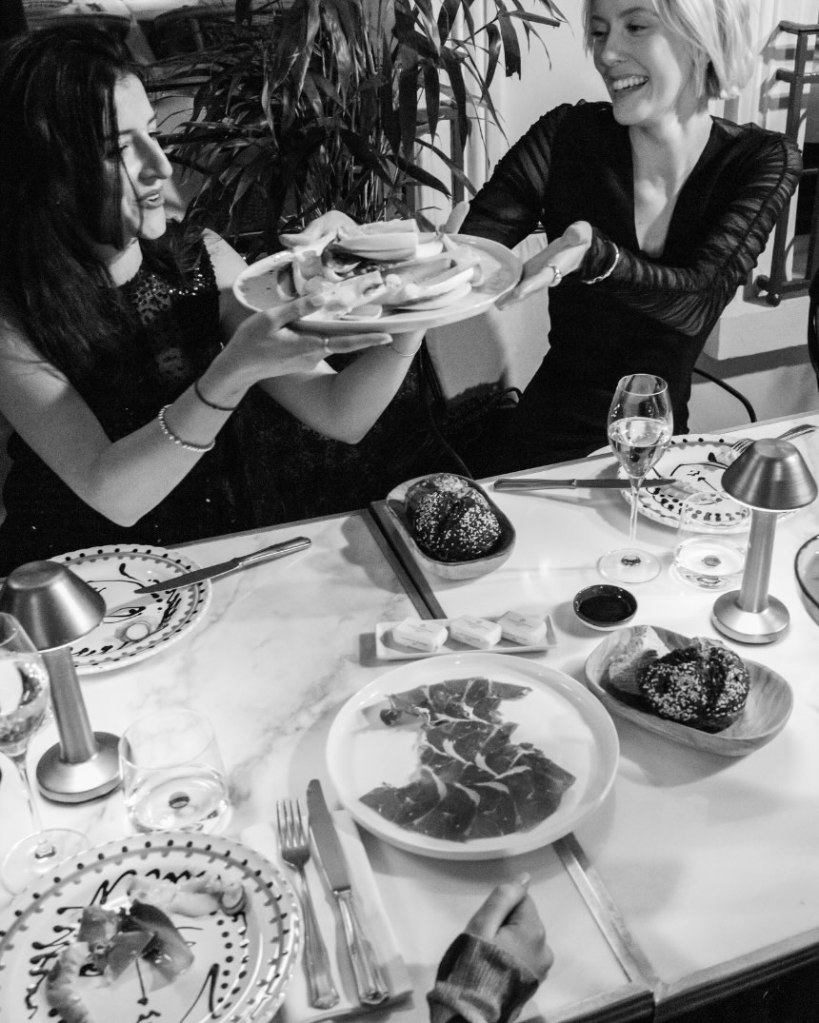 two women sitting at a table full of delicious food, passing plates to one another