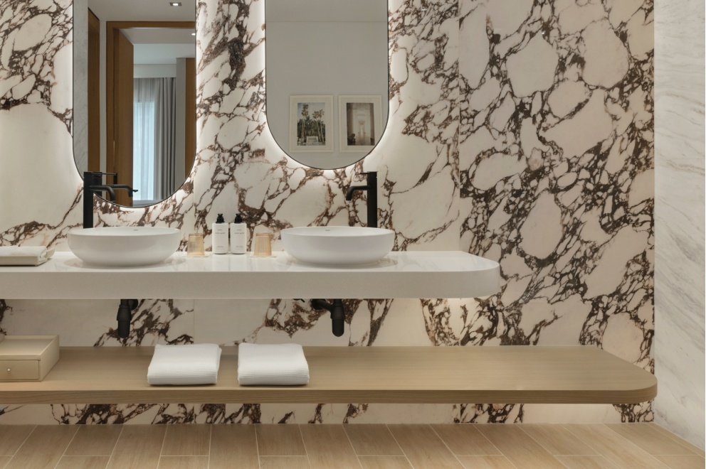 Uniquely marbled bathroom with long oval-shaped mirrors above wash basins with black faucets and a lower shelf with towels