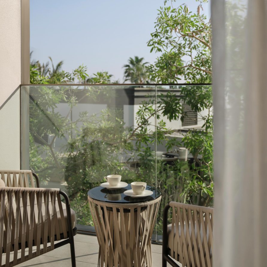 A small table with coffee cups sits between two chairs on a sunny balcony surrounded by leafy greenery at the Delano Hotel, Dubai.