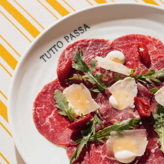 Image of beef carpaccio on a plate that says Tutto Passa atop a yellow and white striped tablecloth