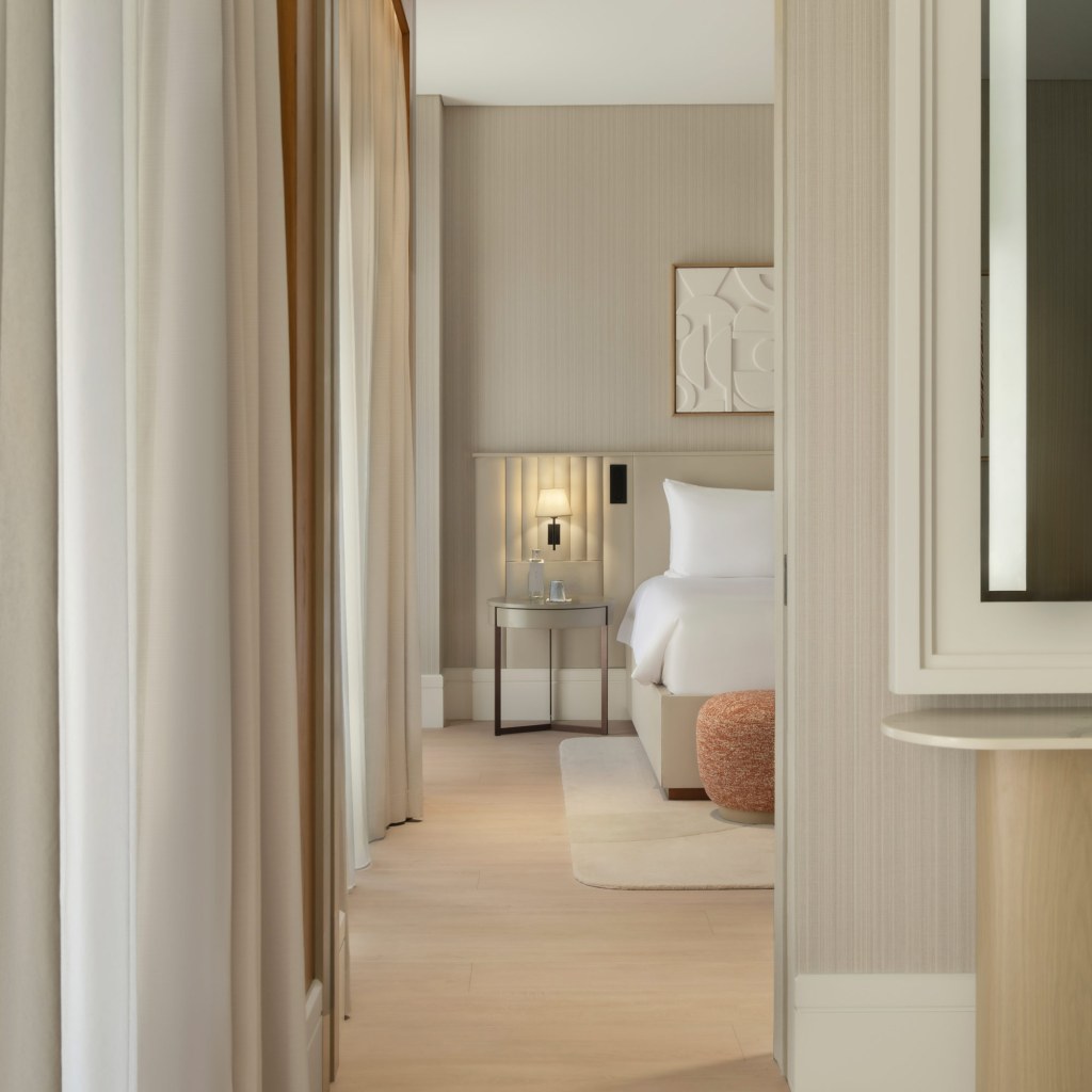 A modern hotel room at Delano Hotel, Dubai, features a neatly made bed beside a nightstand with a lamp. Soft curtains and beige tones enhance the elegant atmosphere.