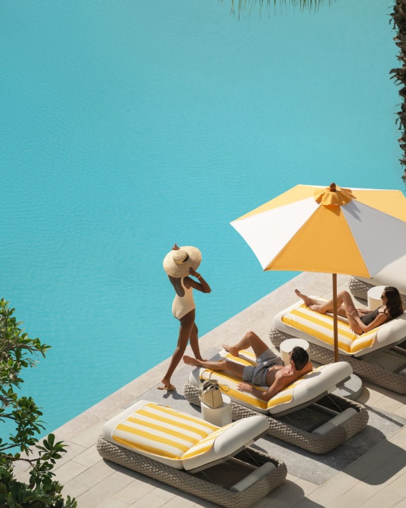 a woman in a yellow swimsuit and a large hat walking by the pool 