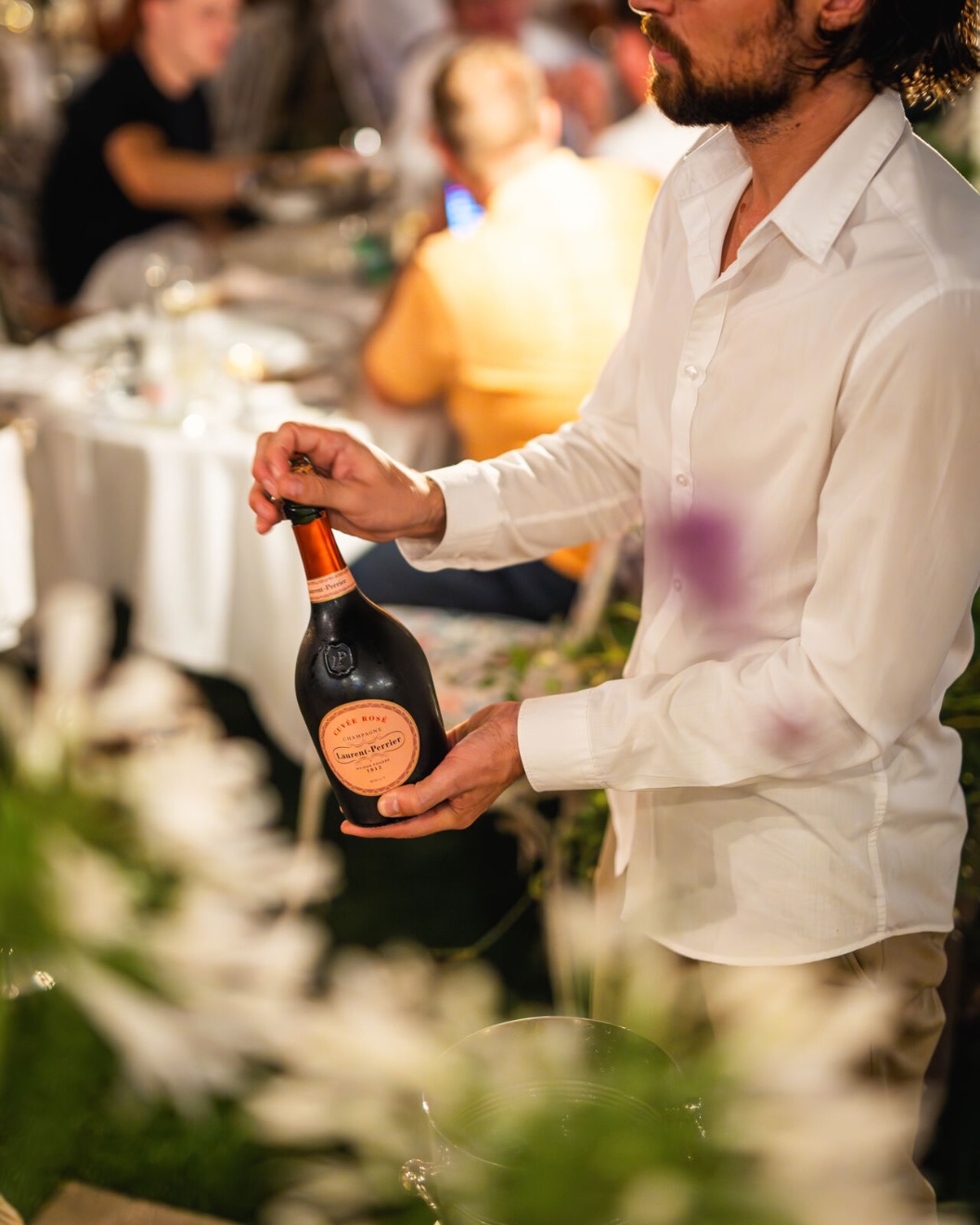 A person opens a champagne bottle at a Delano Hotel event in Dubai, surrounded by elegantly dressed guests seated at a table with white tablecloths under soft lighting.
