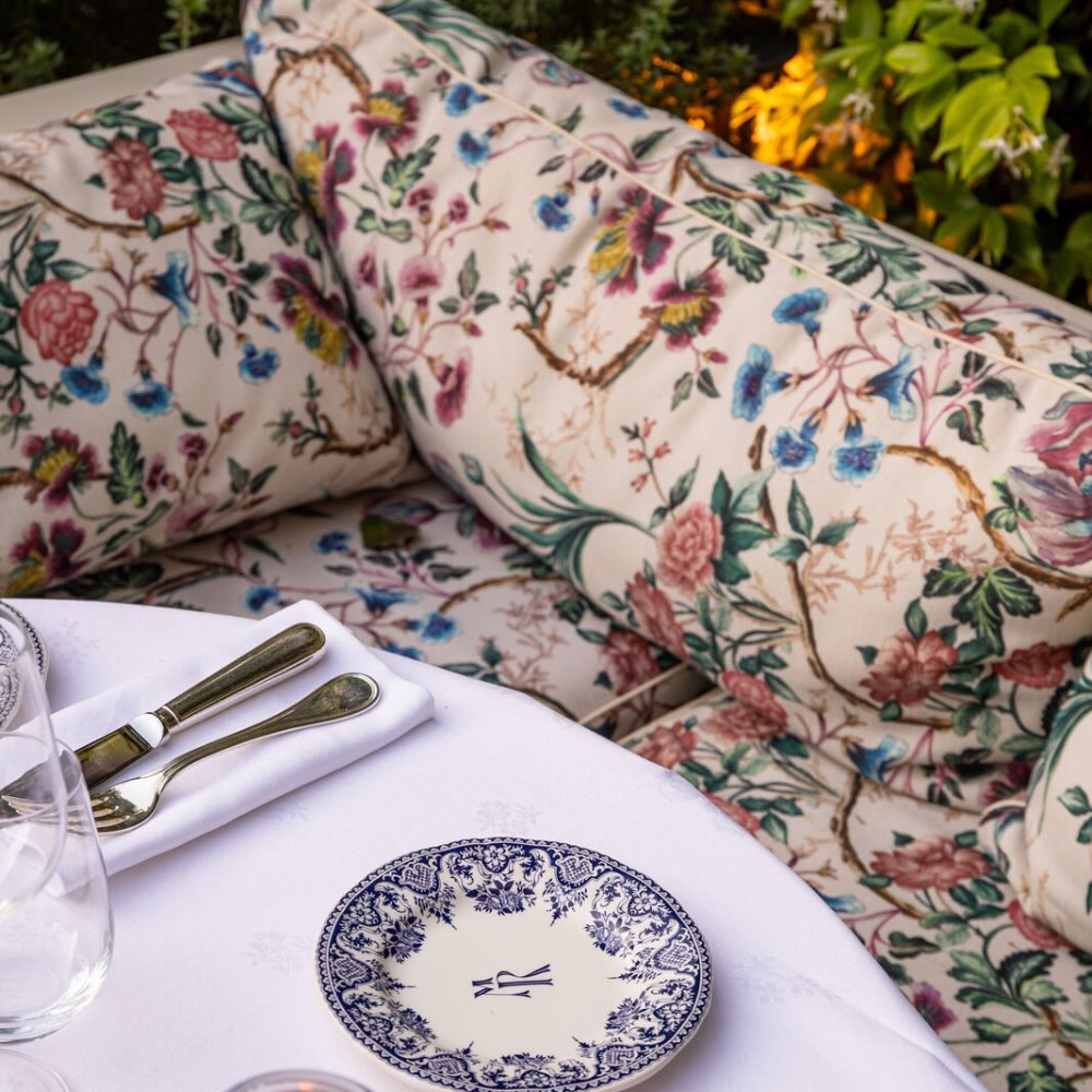 a floral couch as seating for a white linen draped table with a blue and white plan with the MR logo