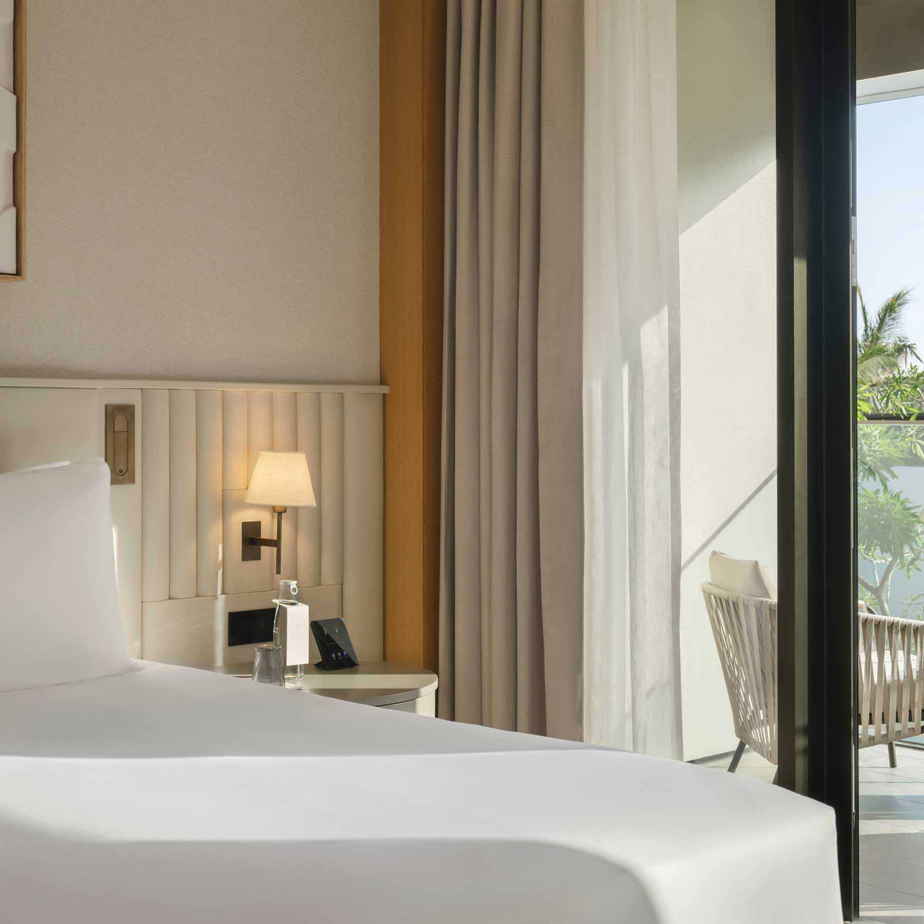 a side view of a king bed with a bedside table and lit up sconce light above it and sliding glass doors that lead to a balcony outside