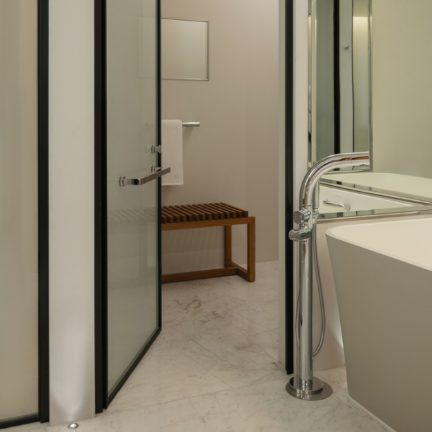A modern bathroom features an elegant freestanding bathtub with sleek fixtures, set against a minimalist design with glass doors and a wooden bench in the Delano Hotel, Dubai.
