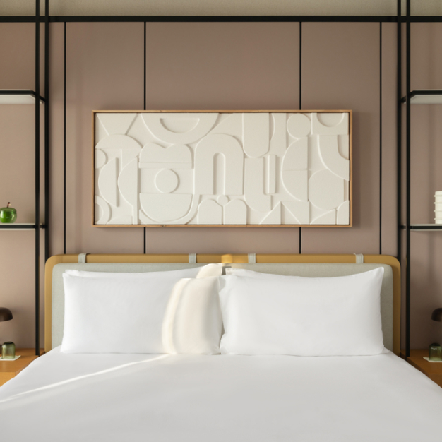 A neatly made bed rests under abstract wall art in a stylish, minimalist room at Delano Hotel, Dubai. The modern decor includes soft tones and geometric shelving.