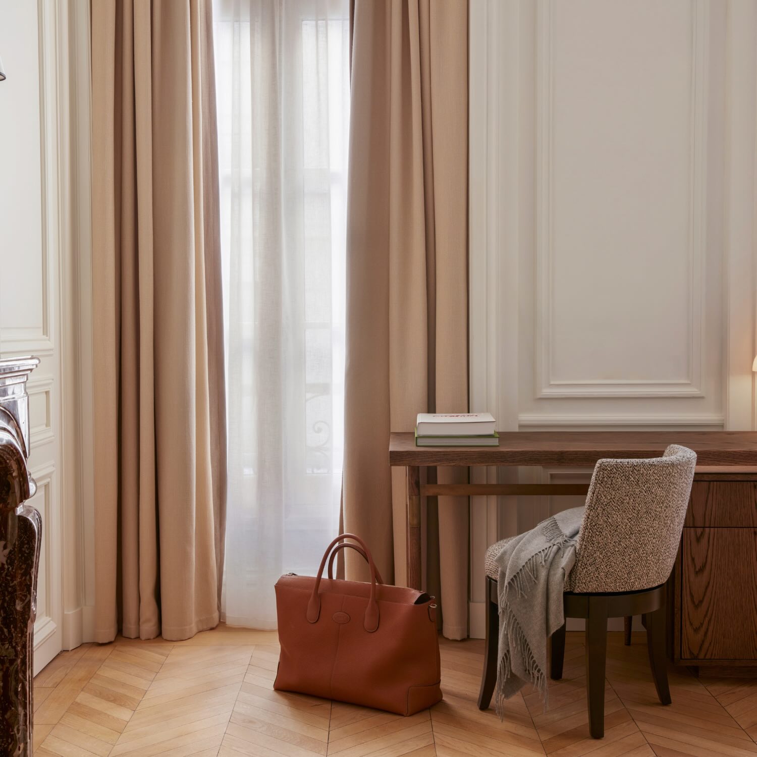 a desk up against a wall next to a window with drapes and a leather bag sitting on the floor next to the chair