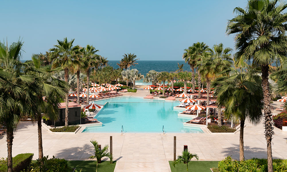 A large pool with rows of loungers and orange umbrellas, surrounded by palm trees.