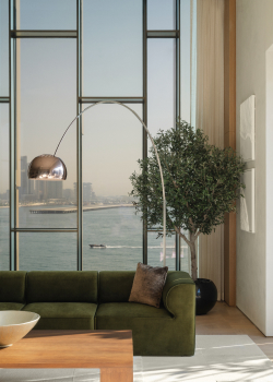 Room with floor to ceiling glass windows overlooking water and the Dubai skyline, with a green sofa and silver arc floorlamp