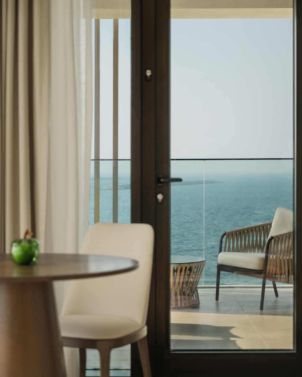 Serene view from an elegant interior featuring a table and chair, looking out through glass doors to a balcony with comfortable seating and a stunning ocean backdrop.