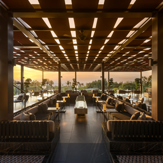 Modern outdoor lounge area with sleek black furniture, warm lighting, and a contemporary wooden ceiling design, overlooking a serene sunset and lush greenery.