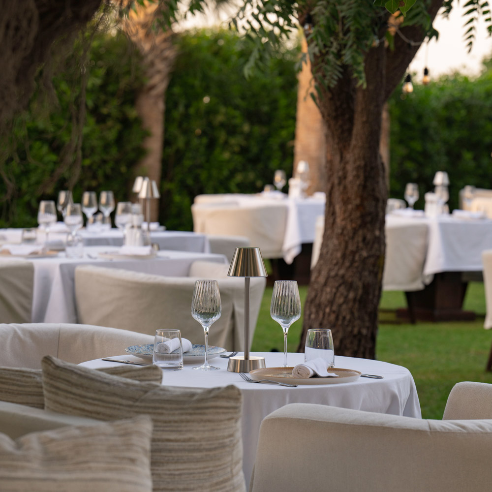 Elegant outdoor dining setup in a lush garden setting, featuring round tables draped with white tablecloths, modern table lamps, wine glasses, and comfortable beige cushioned chairs. Trees and greenery surround the area, creating a relaxed and sophisticated ambiance.