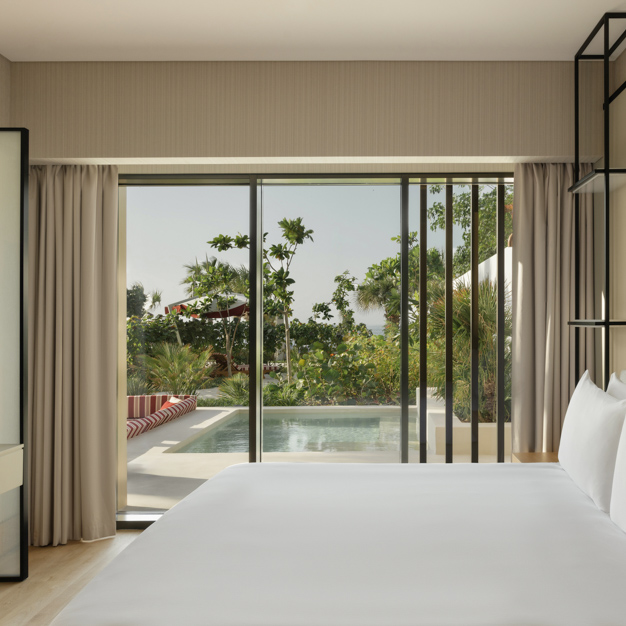 Luxurious hotel room featuring a king-size bed with crisp white linens, and floor-to-ceiling glass doors opening to a private outdoor pool surrounded by lush greenery. Built-in seating with red and white striped cushions is visible by the pool, creating a seamless indoor-outdoor relaxation space.