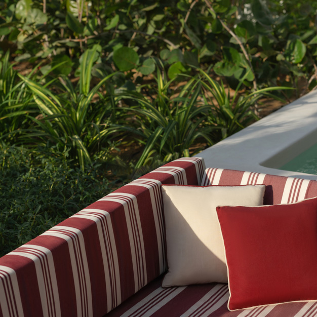 Primer plano de una zona de asientos empotrada en el exterior con cojines a rayas rojas y blancas, acentuados con cojines de color crema y rojo intenso. Los asientos están situados junto a la piscina, rodeados de exuberantes plantas verdes que crean un ambiente acogedor y tropical.