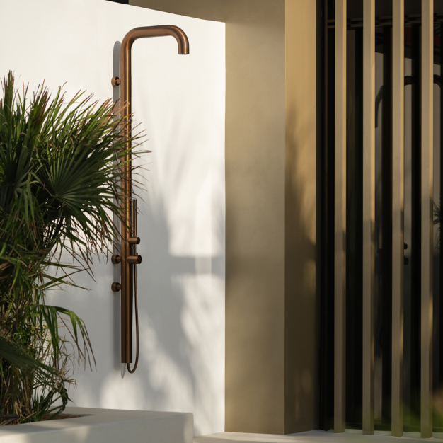 Outdoor shower with a sleek, curved copper fixture mounted on a white wall, surrounded by lush green plants. Adjacent to the shower is a modern architectural structure with vertical slats and beige tones, creating a stylish, minimalist outdoor setting.