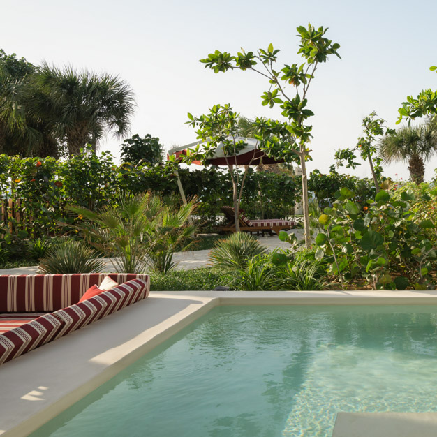 Serene outdoor pool area surrounded by lush greenery and tropical plants. The pool features built-in seating with red and white striped cushions. In the background, wooden sun loungers and a white and burgundy umbrella offer a relaxing spot under the palm trees.