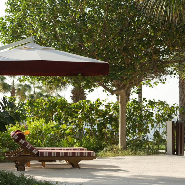 Tranquil outdoor lounge area featuring a wooden sun lounger with red and white striped cushions, positioned under a large white and burgundy patio umbrella. Surrounded by lush greenery and trees, with a view of the sea in the background, creating a relaxed, tropical atmosphere.