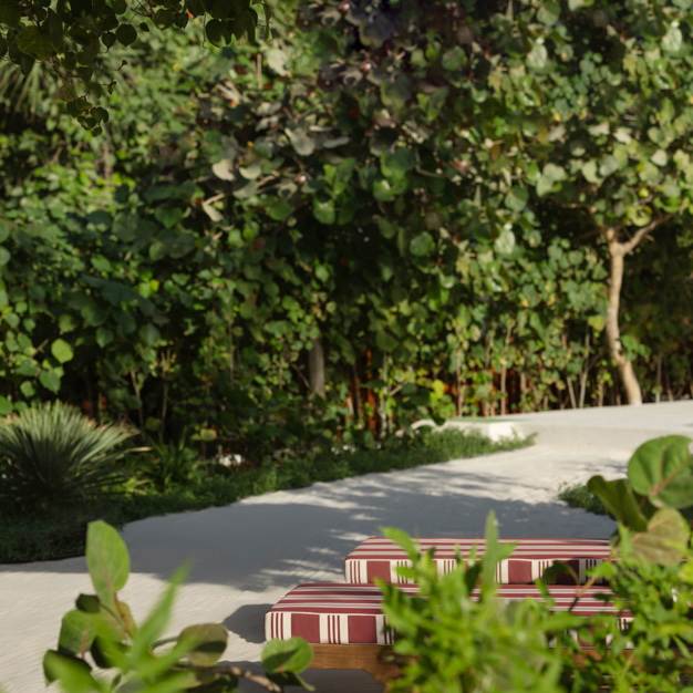 Lush outdoor garden area with a winding sandy pathway surrounded by dense greenery and tropical plants. Red and white striped sun lounger cushions are partially visible in the foreground, blending into the vibrant, natural setting.