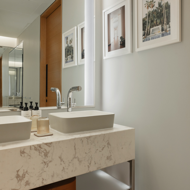 Contemporary bathroom with a white marble countertop featuring two rectangular vessel sinks and sleek chrome faucets. Framed artwork decorates the light grey wall, and a large mirror enhances the sense of space. Modern amenities and subtle lighting create a clean, sophisticated atmosphere.