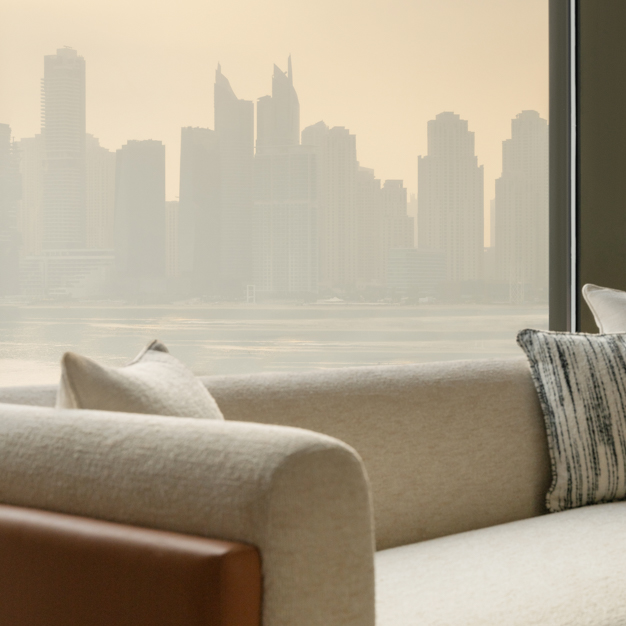 Close-up of a modern sofa with neutral-toned and patterned cushions, positioned next to a large window offering a view of a city skyline with skyscrapers, partially veiled in a soft haze. The scene exudes a tranquil, urban atmosphere.