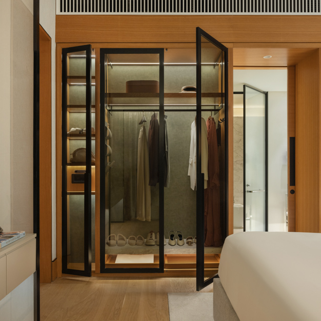 Modern hotel room wardrobe with sleek black-framed glass doors, illuminated interior, and neatly arranged clothing, shoes, and accessories. The open closet is integrated into a warm wood-accented room, with a glimpse of the bathroom and part of a neatly made bed visible in the background.