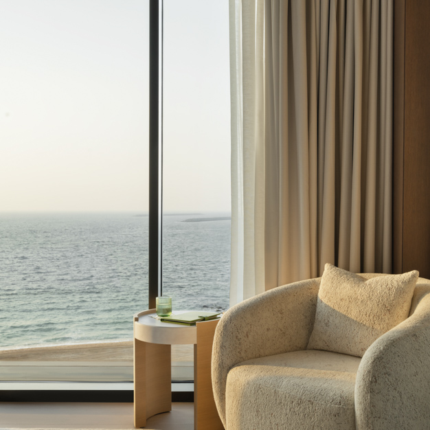 Cozy seating nook by a floor-to-ceiling window with a stunning ocean view. A plush cream-colored armchair is paired with a round wooden side table holding a drink and a book. Soft beige curtains frame the window, creating a calm and relaxing atmosphere.