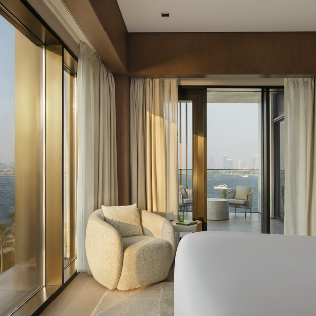 Luxurious hotel bedroom with floor-to-ceiling windows offering panoramic ocean views. A plush cream-colored armchair sits in the corner beside light beige curtains, while a king-size bed is partially visible. A private balcony with seating overlooks the water, with boats and a city skyline in the distance.