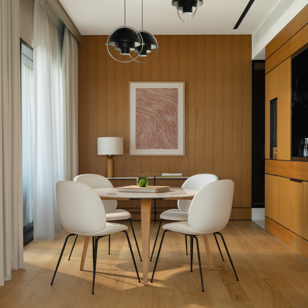 Contemporary dining area in a hotel suite, featuring a round wooden table surrounded by four white chairs with black legs. Pendant lights hang from the ceiling, while a floor lamp and abstract artwork decorate a wood-paneled accent wall. Floor-to-ceiling curtains and built-in cabinetry enhance the modern, warm ambiance.
