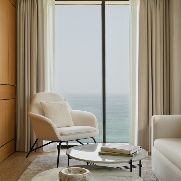Cozy seating area in a modern hotel room, featuring a white armchair with a cushion, a round marble coffee table with books and decor, and floor-to-ceiling windows offering a stunning ocean view. Soft beige curtains frame the windows, enhancing the serene and elegant ambiance.