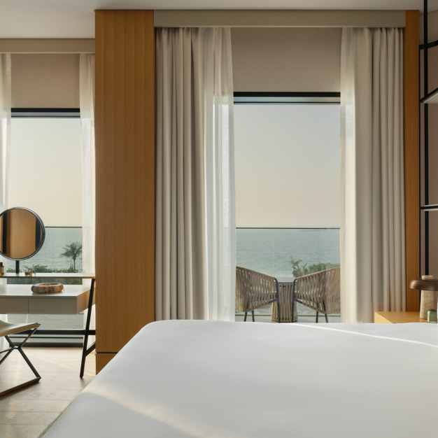Modern hotel room with a king-size bed, a vanity desk with a round mirror, and floor-to-ceiling windows offering a view of the ocean and palm trees. A balcony with two chairs and a table overlooks the sea, while soft curtains and wooden accents create a calming, elegant atmosphere.