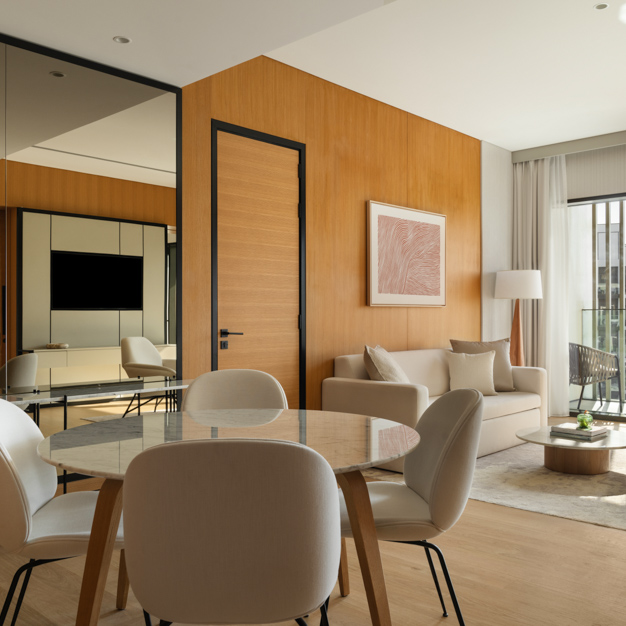 Contemporary hotel suite with an open-plan living and dining area, featuring a round marble dining table with four white chairs, a beige sofa with cushions, a wooden coffee table, and a floor lamp. Large windows with curtains let in natural light, and a mirror wall reflects the modern interior design.