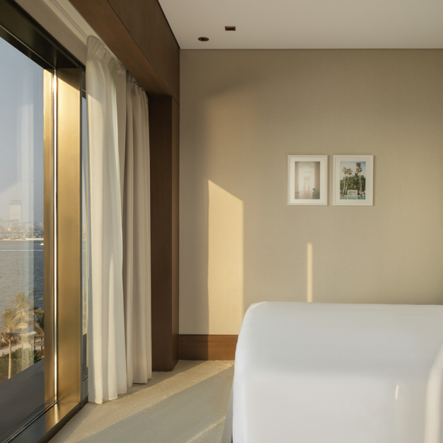 Minimalist hotel bedroom with a king-size bed, white linens, and floor-to-ceiling windows offering an ocean view. Framed artwork hangs on a neutral-toned wall, with sunlight streaming into the room.