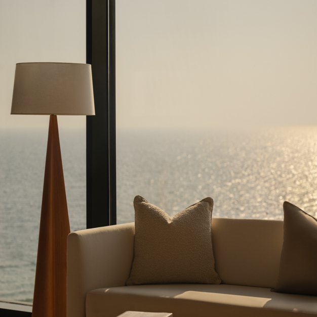 Close-up of a hotel room corner with a cream sofa, a wooden floor lamp, and a large window overlooking the sunlit ocean, creating a tranquil seaside ambiance.