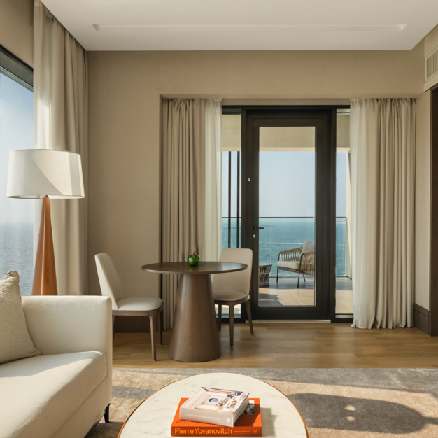 Hotel suite’s living area featuring a round dining table, two chairs, a floor lamp, and a cream sofa, with floor-to-ceiling windows and a balcony door offering panoramic ocean views.