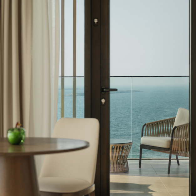 View from inside a hotel room looking through a glass door onto a balcony with woven seating, overlooking the ocean. A small dining area with a round wooden table and a cream chair is visible inside.