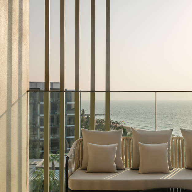 Cozy balcony nook with a woven bench-style seat and cream cushions, offering a view of the ocean and neighboring buildings. Vertical slats provide partial privacy while maintaining an airy feel.