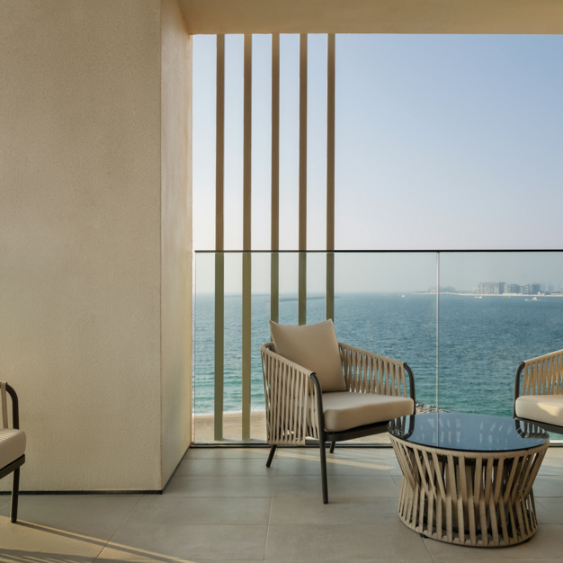 Private balcony with two woven armchairs and a glass-topped coffee table, overlooking the ocean. Vertical slats partially frame the view, enhancing the modern, open-air design.