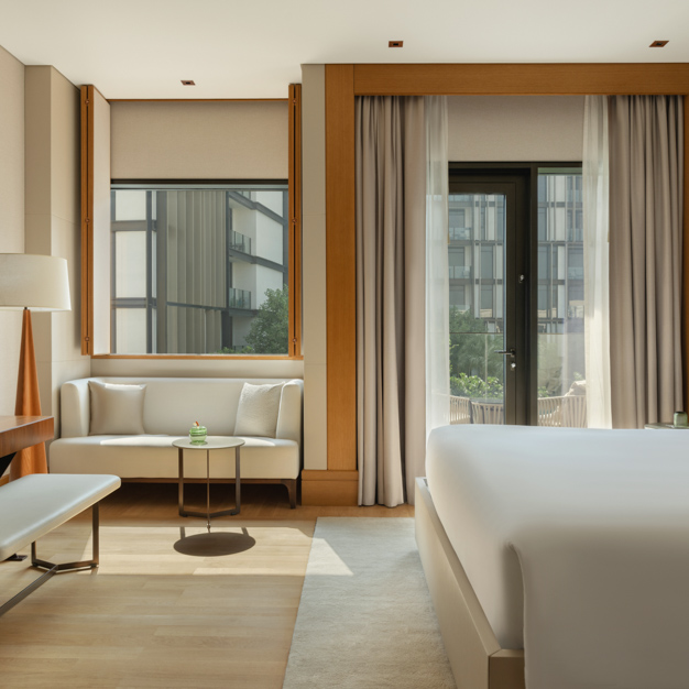 Bright hotel room featuring a king-size bed, a cozy seating nook with a white sofa, a round coffee table, and a desk with a bench. Floor-to-ceiling curtains frame a glass door leading to a private balcony overlooking the hotel’s courtyard.