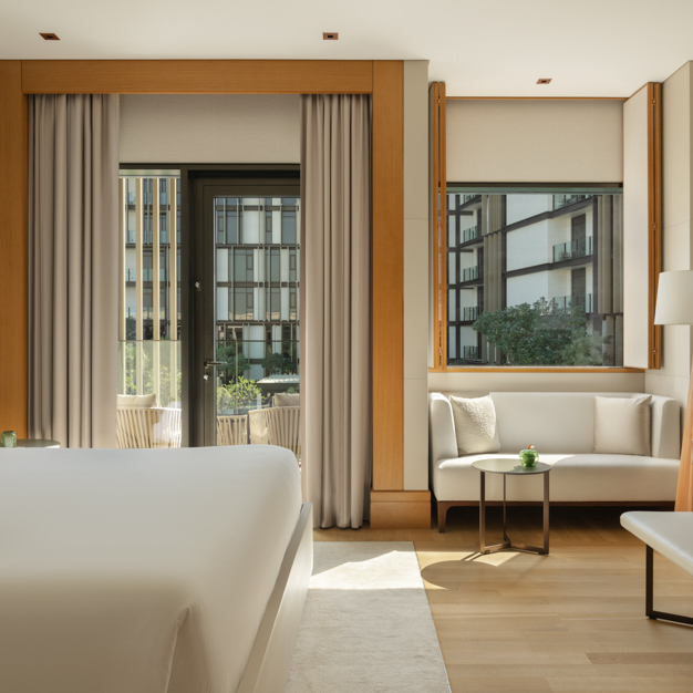 Modern hotel room with a king-size bed, a white sofa seating area, and a floor-to-ceiling glass door leading to a balcony with outdoor seating. Large windows let in natural light and offer views of the hotel's courtyard and surrounding buildings.