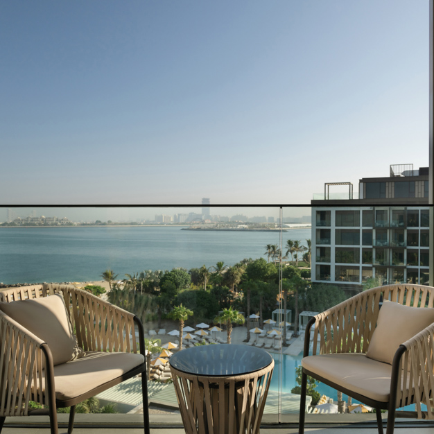 Scenic balcony view overlooking a resort pool, palm trees, and the ocean, with a modern city skyline in the distance. Two woven chairs with cushions and a small round table provide a relaxing outdoor seating area with glass railing enhancing the view.