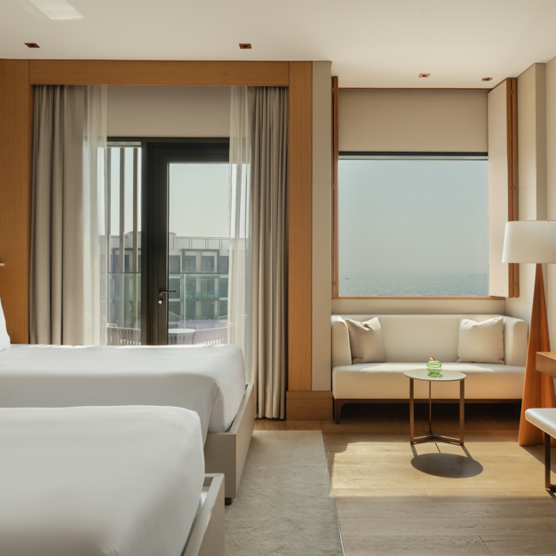 Modern hotel room featuring two twin beds, a small seating area with a white sofa and coffee table, a floor lamp, and floor-to-ceiling windows with a glass door leading to a balcony, overlooking the sea and a neighboring building.