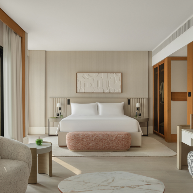 Elegant hotel bedroom featuring a king-size bed with crisp white linens, a textured pink bench at the foot, and modern bedside tables with matching lamps. A built-in open wardrobe with wooden accents is visible, alongside a seating area with a round marble coffee table. Neutral tones, soft textures, and natural light create a calming, sophisticated atmosphere.