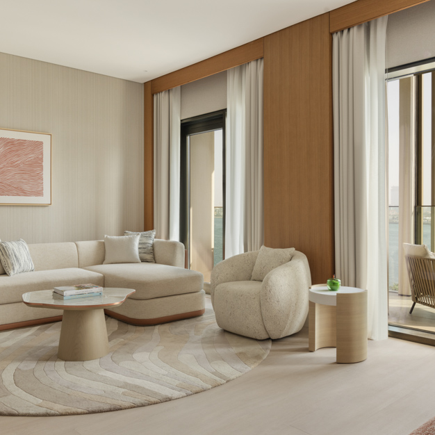 Modern living area in a hotel suite with a curved cream sofa, a plush armchair, and a round coffee table on a textured area rug. Floor-to-ceiling windows with light curtains offer ocean views and lead to a balcony with outdoor seating. Wood panel accents and neutral tones create a warm, inviting atmosphere.