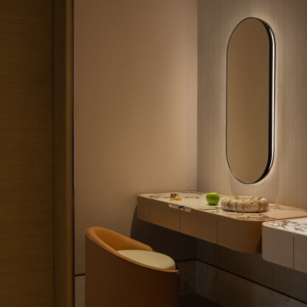 Sophisticated vanity area featuring a marble countertop with a curved design, a built-in desk, and a mustard-colored upholstered chair. An oval mirror with soft backlighting is mounted on a textured beige wall, creating a warm, elegant ambiance. Small decorative items and a drink are placed on the counter.