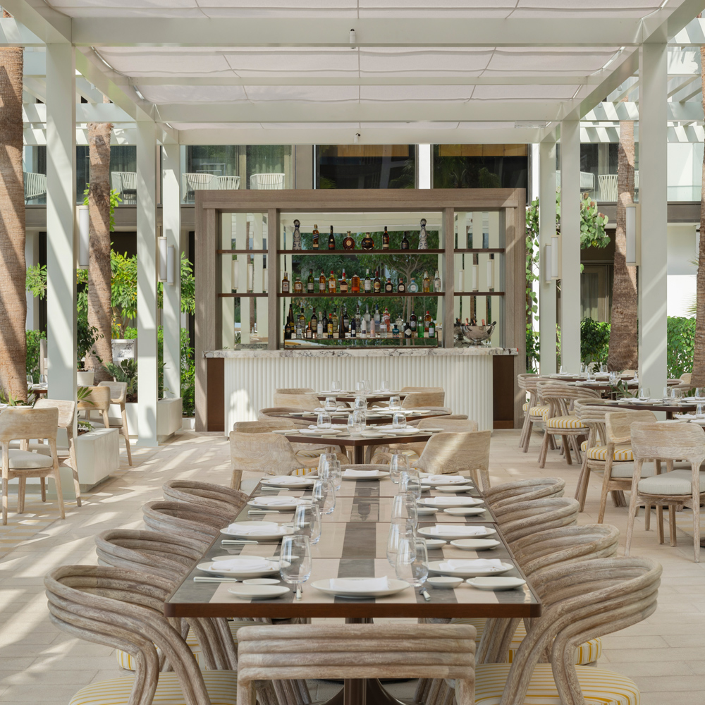 A stylish open-air restaurant with elegant wooden dining tables set with white plates and glassware. The space is bathed in natural light, featuring a chic bar with a curated selection of beverages, lush greenery, and a sophisticated ambiance perfect for dining and social gatherings.