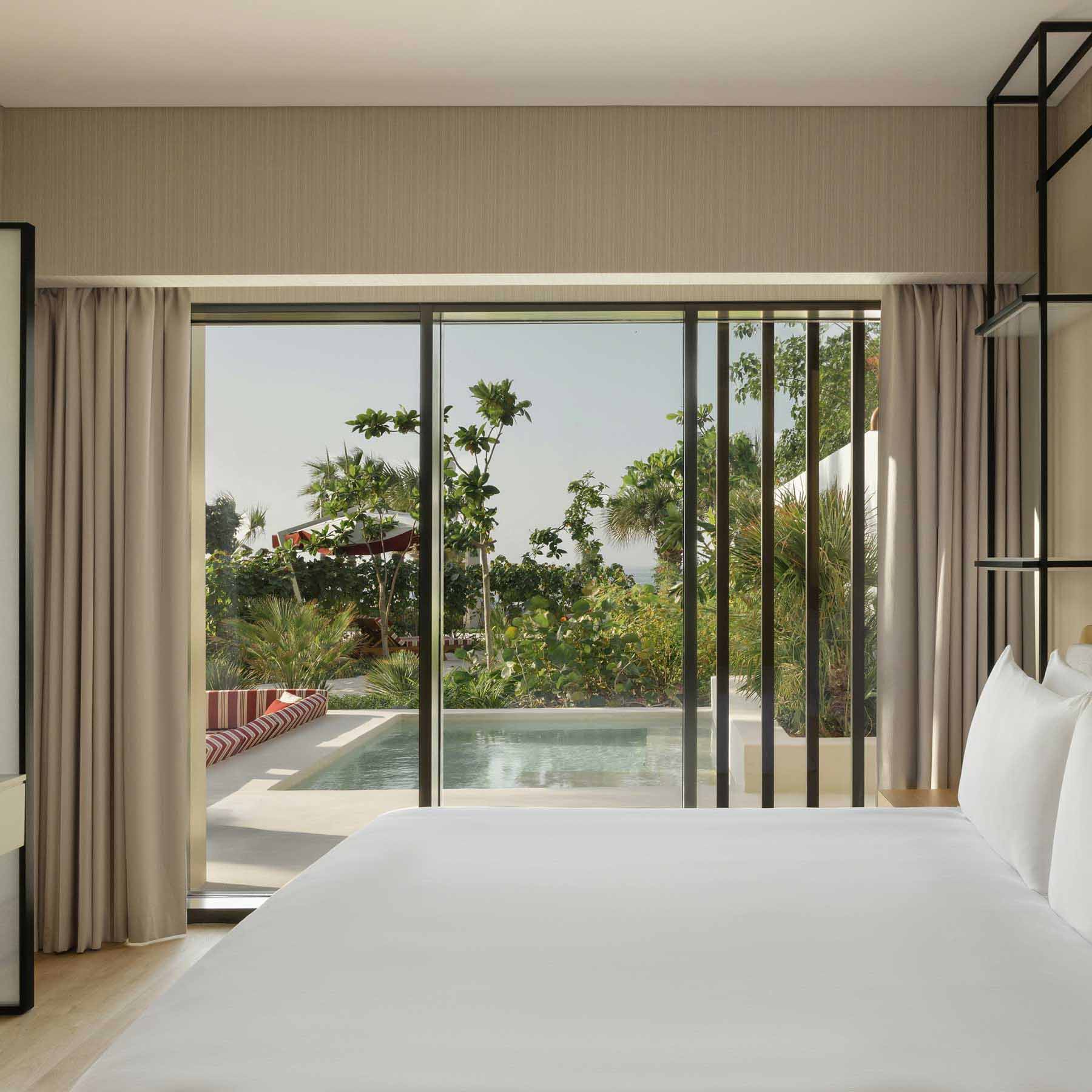 Luxurious hotel room featuring a king-size bed with crisp white linens, and floor-to-ceiling glass doors opening to a private outdoor pool surrounded by lush greenery. Built-in seating with red and white striped cushions is visible by the pool, creating a seamless indoor-outdoor relaxation space.