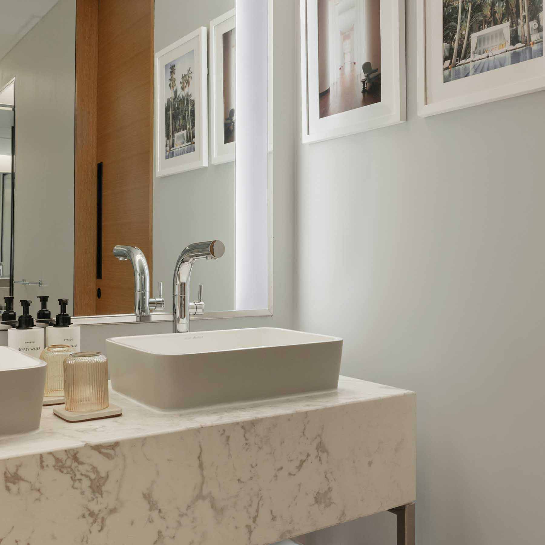 Contemporary bathroom with a white marble countertop featuring two rectangular vessel sinks and sleek chrome faucets. Framed artwork decorates the light grey wall, and a large mirror enhances the sense of space. Modern amenities and subtle lighting create a clean, sophisticated atmosphere.