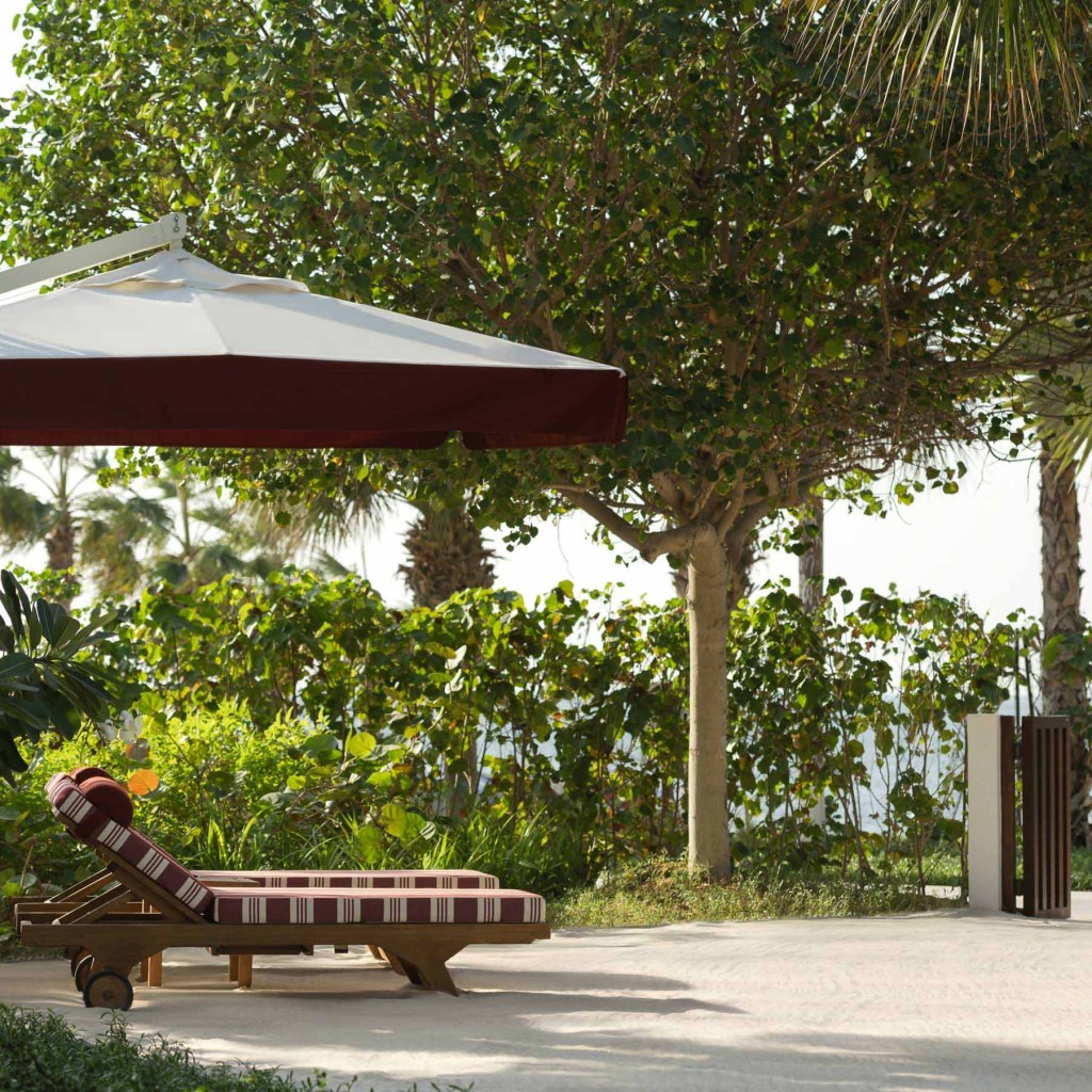 Tranquil outdoor lounge area featuring a wooden sun lounger with red and white striped cushions, positioned under a large white and burgundy patio umbrella. Surrounded by lush greenery and trees, with a view of the sea in the background, creating a relaxed, tropical atmosphere.