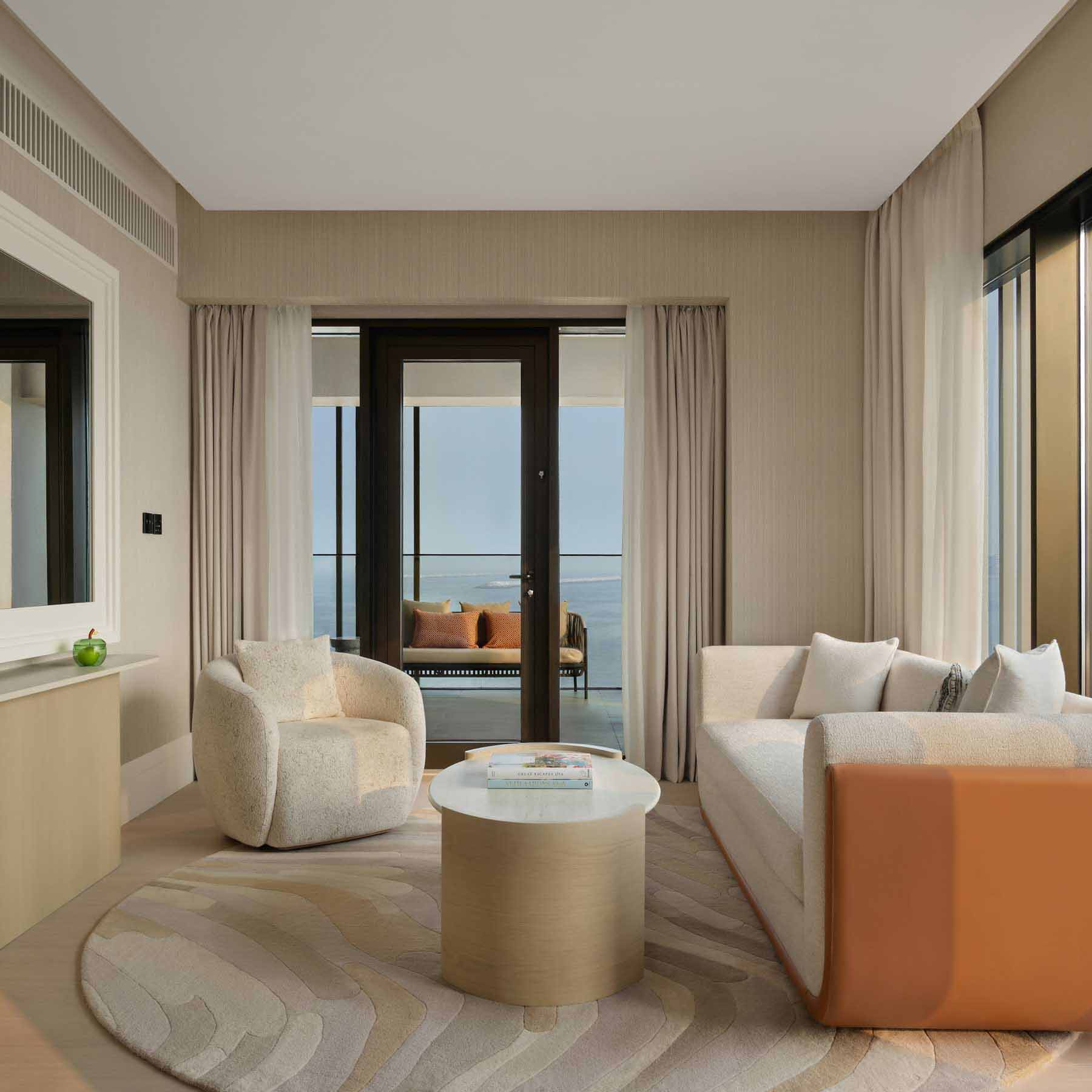 Stylish hotel suite living room featuring a curved white and tan sofa, a cozy cream armchair, and a round marble coffee table on a patterned area rug. Floor-to-ceiling glass doors lead to a balcony with ocean views and additional seating, while neutral tones and soft textures create a relaxed, elegant atmosphere.